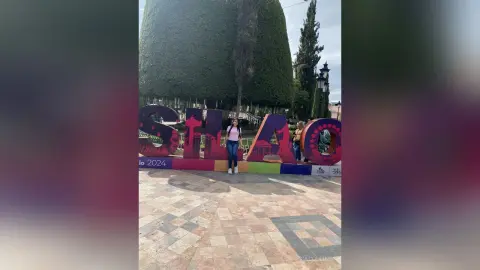 Young woman in a white top and jeans standing by colorful giant letters in a sunny outdoor plaza.