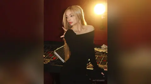 Elegant blonde woman in a black off-the-shoulder dress posing at a casino gaming table under warm lighting.