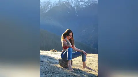 Thoughtful woman in a red tank top and blue jeans sitting on a grassy hill against a misty mountain landscape.