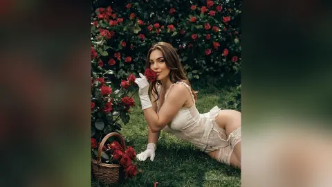 Elegant woman in white lace lingerie holding a red rose in a romantic rose garden setting.