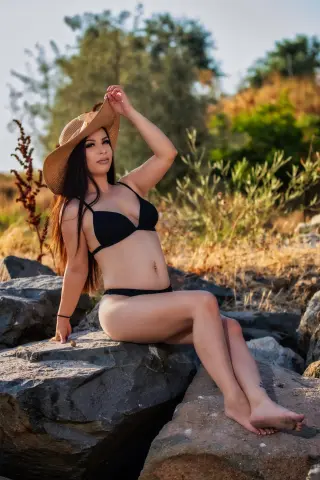 Woman in black bikini and sun hat sitting on rocks in a sunny outdoor landscape.