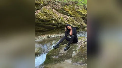 Young woman in black long-sleeve outfit sitting on rocks by a forest stream in a peaceful natural setting.