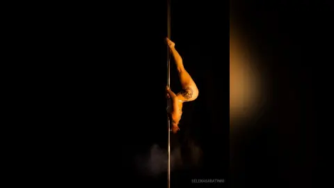 Athletic woman in a white bikini performing a graceful pole dance move in a moody, dark studio setting.