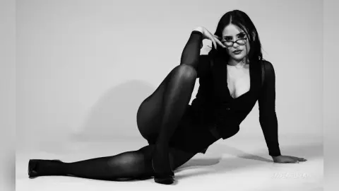 Stylish woman in black outfit and glasses posing elegantly in a moody black and white studio portrait.