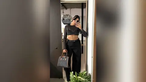 KimberlyIsaza poses in a black long-sleeve crop top and black trousers, holding a patterned handbag in a mirror selfie.