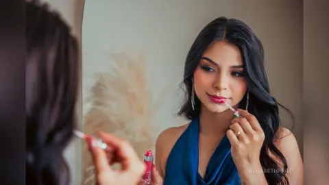 Young woman in a blue halter dress applying pink lipstick in a mirror for a glamorous beauty routine.