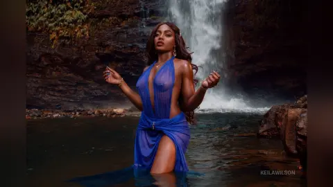 Elegant woman in a flowing blue gown standing in a misty waterfall forest setting with a dreamy mood.