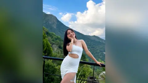 Smiling woman in a white summer dress posing on a balcony overlooking lush green mountains under a blue sky.