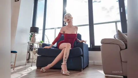 DaisyLin sits on a blue armchair wearing a red off-the-shoulder dress and white strappy heels.