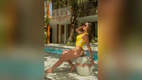 Stylish woman in a bright yellow one-piece swimsuit posing on a white stool by a sunny tropical resort pool.