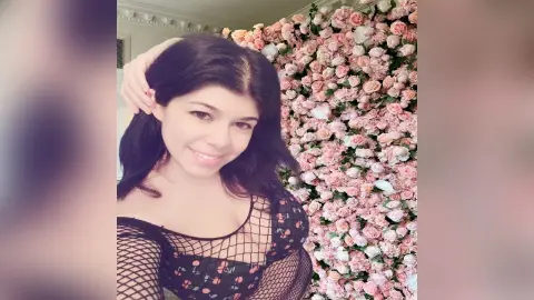 Smiling woman in a black fishnet top posing against a romantic pink floral flower wall background.