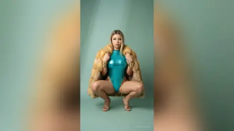 NicolePeach squats in a shiny blue bodysuit and a tan fur coat, showing off her legs and gold bracelets against a plain teal background.