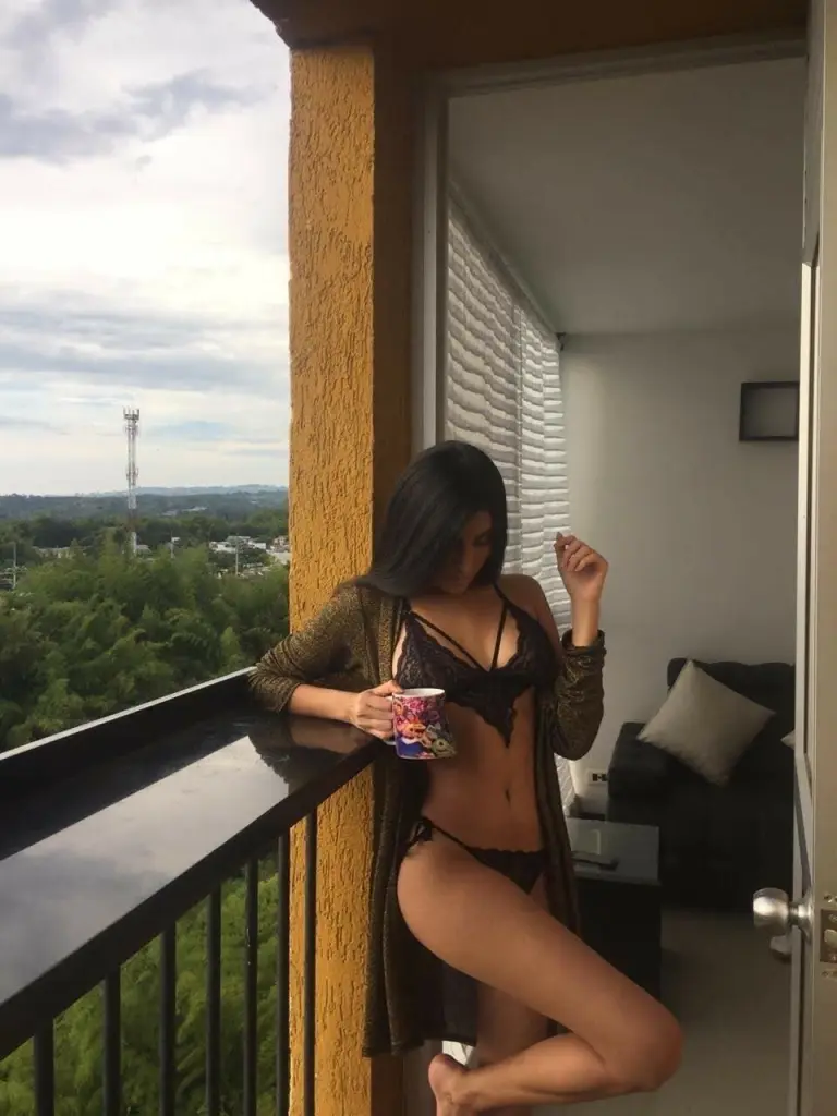 AriaMatheuss poses with her dark hair down, wearing a black bra and a red jacket against a dark night city background.