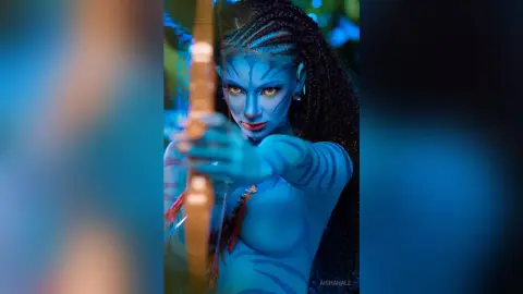 Blue Na'vi female character with braided hair aiming a wooden bow in a mystical forest setting.