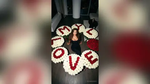 ReynaGomes poses in a black bodysuit surrounded by large white and red flower arrangements spelling out the word LOVE.