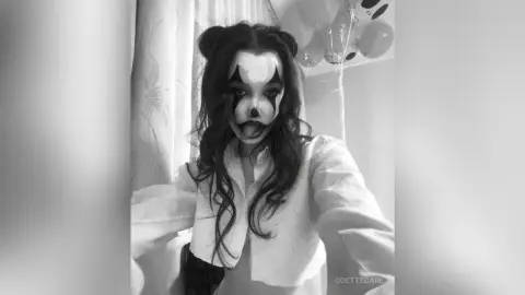 Young woman in spooky clown makeup and white shirt posing in a room with balloons, moody black and white aesthetic.