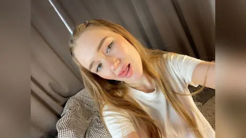 AddieRunk pouts with her tongue out, wearing a white t-shirt and letting her long blonde hair hang down.
