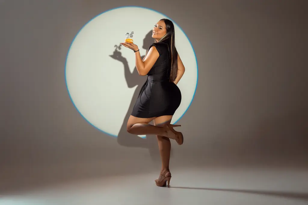 Mia_Laurent smiles while wearing a black sleeveless top, holding a small dessert or treat in her hand against a light grey background.