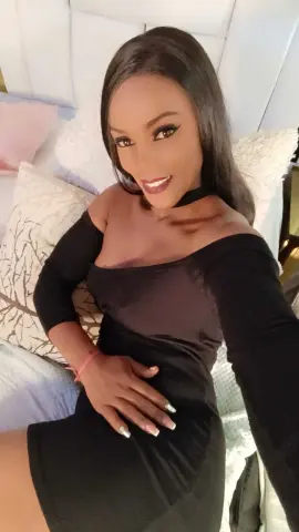 Smiling woman in a chic black long-sleeve dress relaxing on a white bed in a bright bedroom.