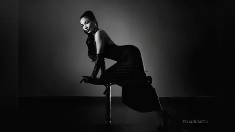 ElliaRussell poses in a black dress and long gloves while sitting on a tall stool against a dark background.