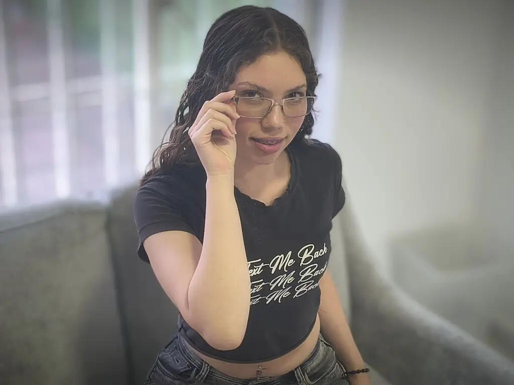 Smiling young woman with wavy hair wearing glasses and a black graphic t-shirt in a bright indoor setting.