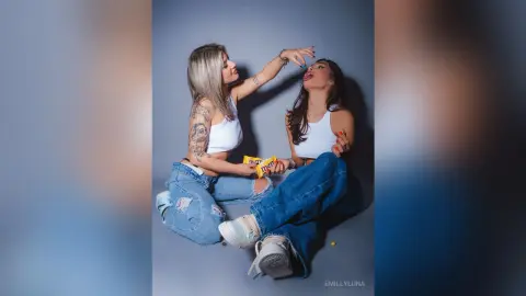Two young women in white crop tops and denim jeans laughing together in a playful studio setting.