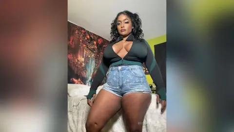 BossyBella — BossyBella poses in a black long-sleeve crop top and denim shorts, showing off her curves against a decorative wall background.