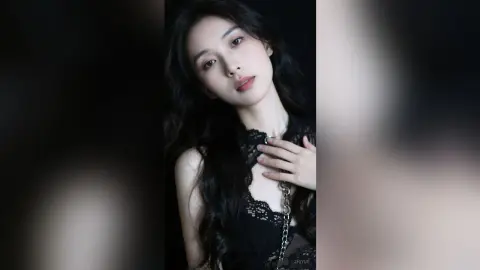 ZhiYue poses with long black hair, wearing a black lace top while looking directly at the camera against a dark background.