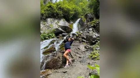Young woman in casual athletic wear standing by a scenic forest waterfall in a lush mountain setting.