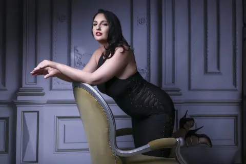 Elegant woman in a black evening dress sitting on a vintage chair against a classic paneled wall background.