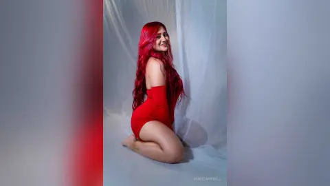 Smiling woman with long red hair wearing a red bodysuit in a bright studio setting.