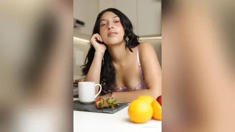 Young woman in a lilac lace bralette relaxing in a bright kitchen with fresh fruit on the table.