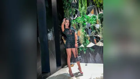 NicoleBella stands in a black tiger-print mini dress, posing against a wall of green leaves and mirrors.
