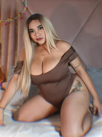 Blonde woman in a brown bodysuit sitting on a bed in a cozy, dimly lit bedroom setting.