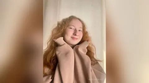 Redheaded woman in a cozy beige sweater smiling in a bright, minimalist indoor setting.