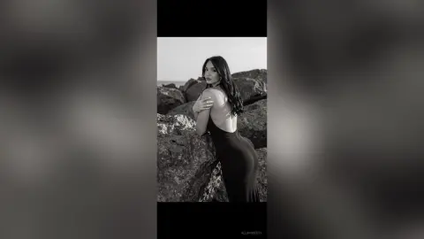 Elegant woman in a black backless dress posing against a rocky coastal landscape in a moody black and white photo.