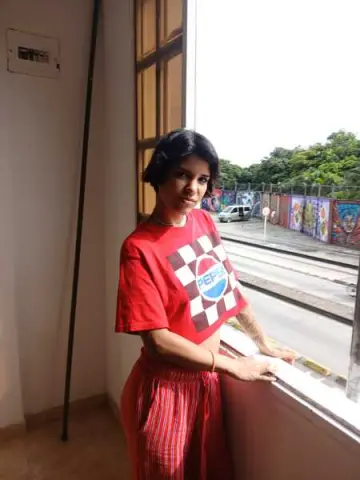 Young woman in a red graphic t-shirt and striped skirt standing by a window in bright daylight.