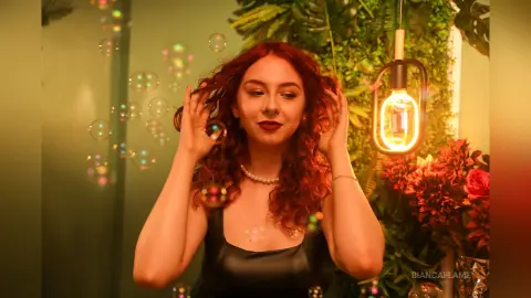 Redheaded woman in a black tank top surrounded by bubbles and warm lighting in a lush indoor garden setting.