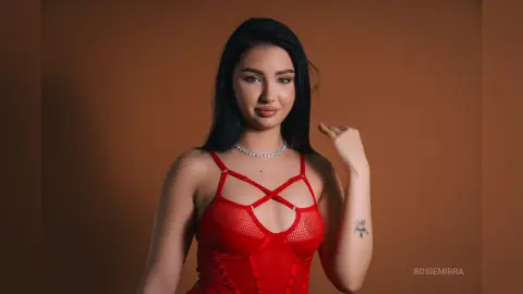 Beautiful young woman in a red mesh bodysuit posing confidently against a warm brown studio background.