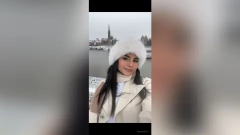 Smiling woman in a white fuzzy hat and beige coat enjoying a snowy winter day by a lake and old town.