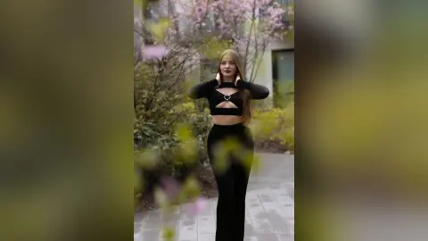 Blonde woman in a black long-sleeve crop top and leggings posing in a dreamy spring garden setting.