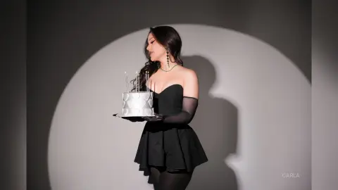 Elegant woman in a black cocktail dress holding a white birthday cake with candles in a moody studio setting.