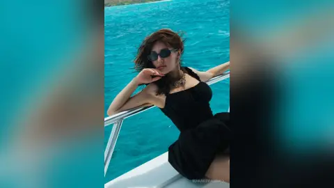 Stylish woman in a black dress and sunglasses relaxing on a boat against a bright blue ocean background.