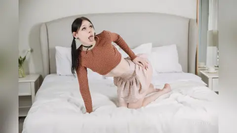 Young woman in a brown striped crop top and pink skirt posing playfully on a white bed in a bright bedroom.