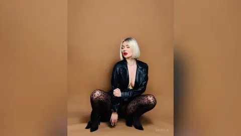 Blonde woman in a black leather jacket and leopard print tights posing against a tan studio background.