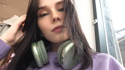 Young woman with long dark hair wearing lavender sweatshirt and sage green headphones in a bright indoor setting.