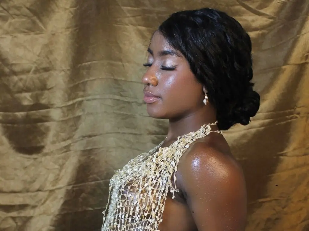 Elegant Black woman in a gold mesh dress posing against a textured bronze backdrop with a serene mood.