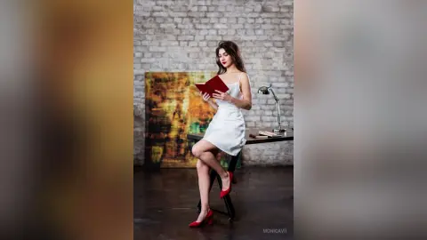 Elegant woman in a white cocktail dress reading a red book in a rustic brick studio setting.