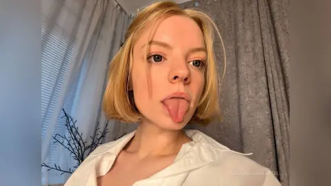 Blonde woman in a white button-down shirt sticking her tongue out in a bright, indoor setting.