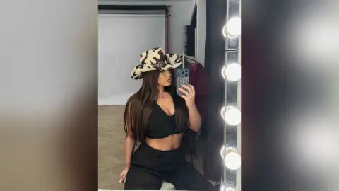 Stylish woman in a black crop top and camo hat posing in a bright studio mirror.